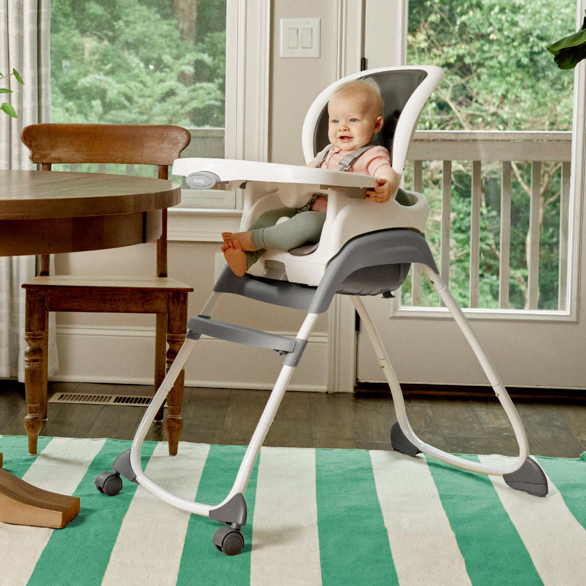 Baby in a high chair in a home setting with a table and windows in the background.
