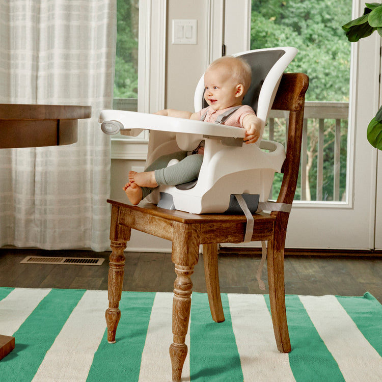 Baby sitting in a white high chair on a wooden chair with a striped rug and window in the background