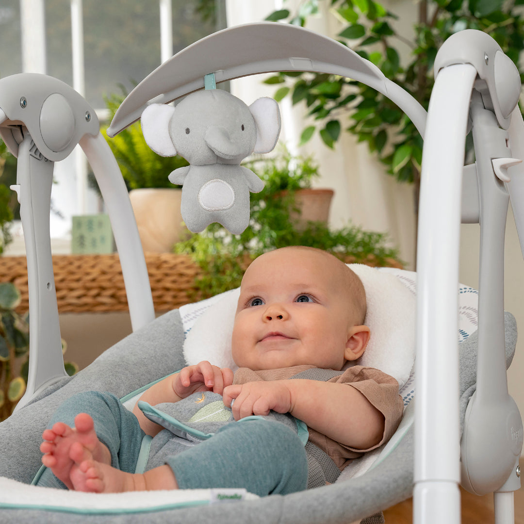 Baby in a gray baby swing with elephant toy, surrounded by greenery