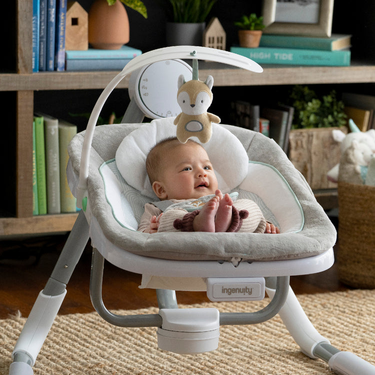 Baby in a white and gray baby swing with a bookshelf in the background