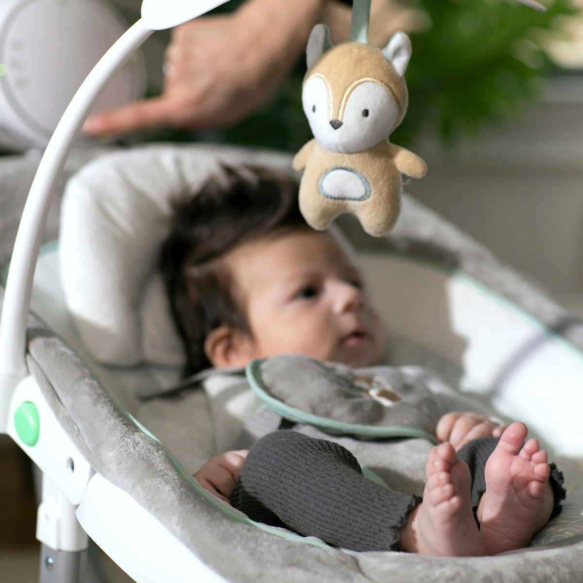 Baby in a white baby bouncer with a plush toy above, blurred background