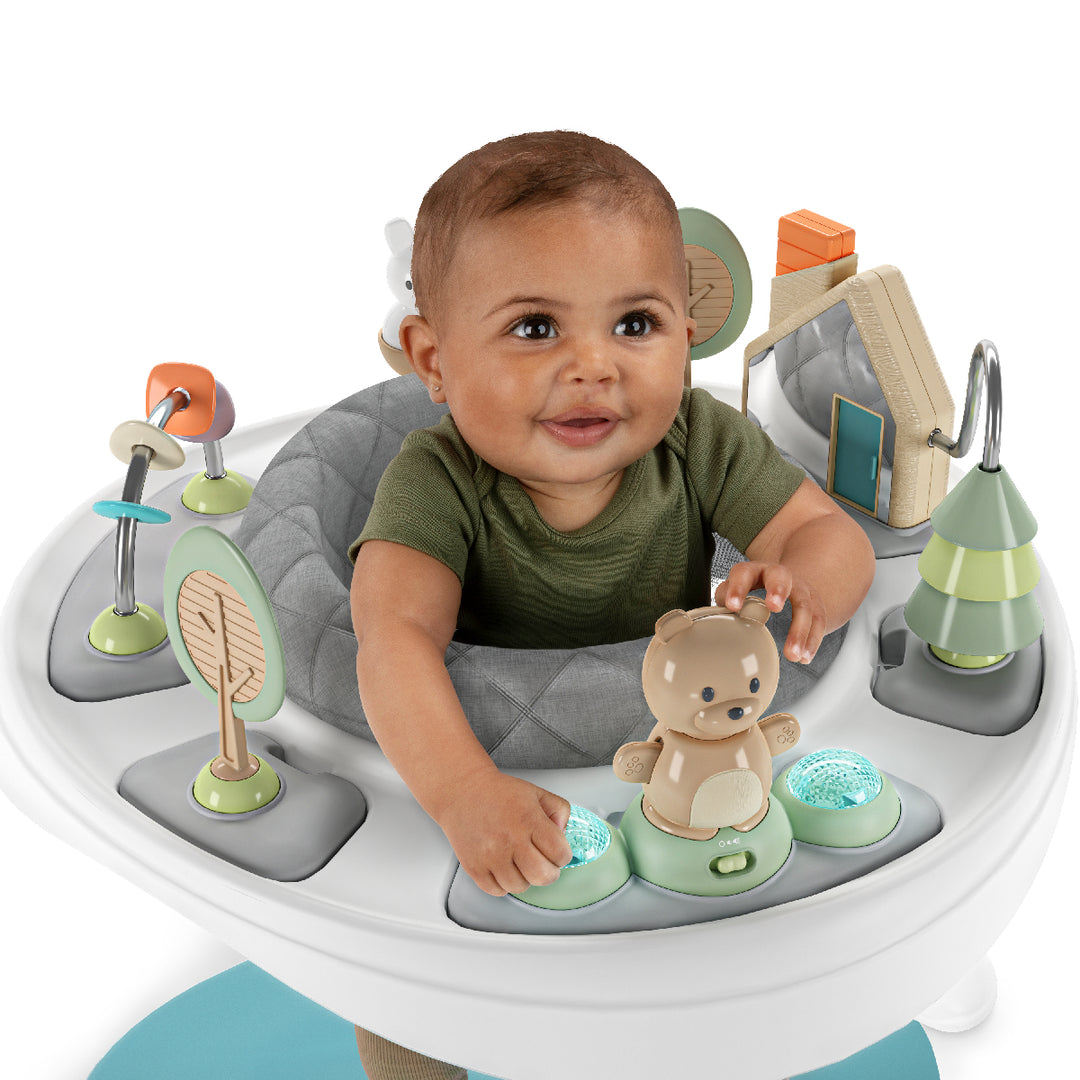 Baby sitting in a gray activity seat with various toys and a teddy bear.