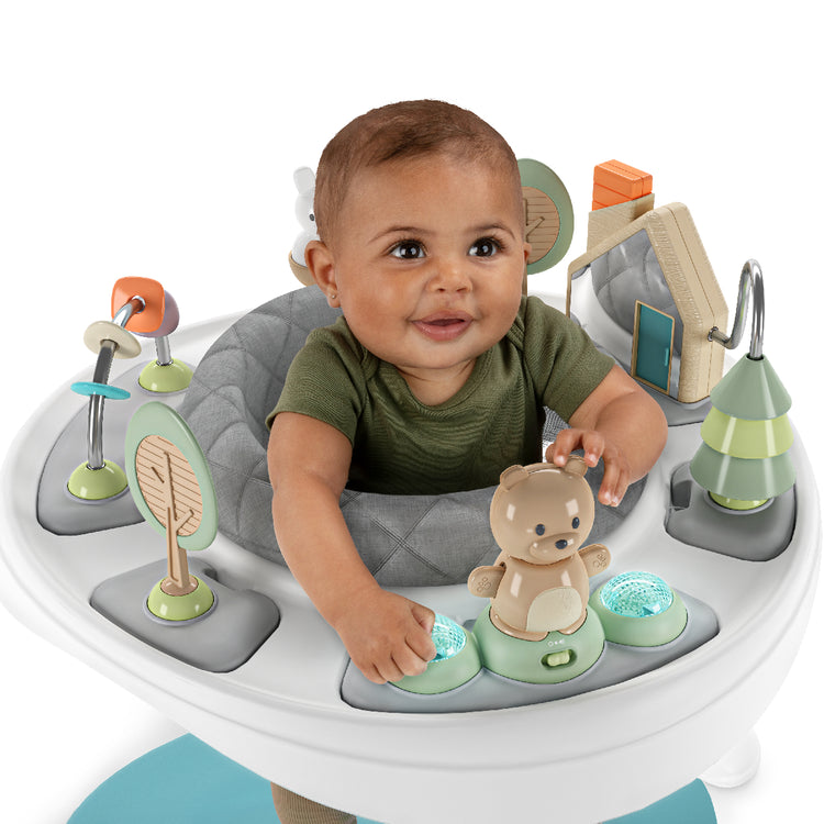 Baby sitting in a gray activity seat with various toys and a teddy bear.