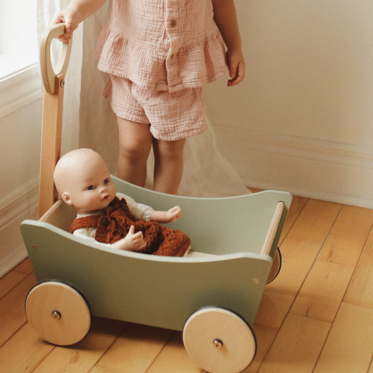 Child with a green toy cart holding a baby doll on a wooden floor.