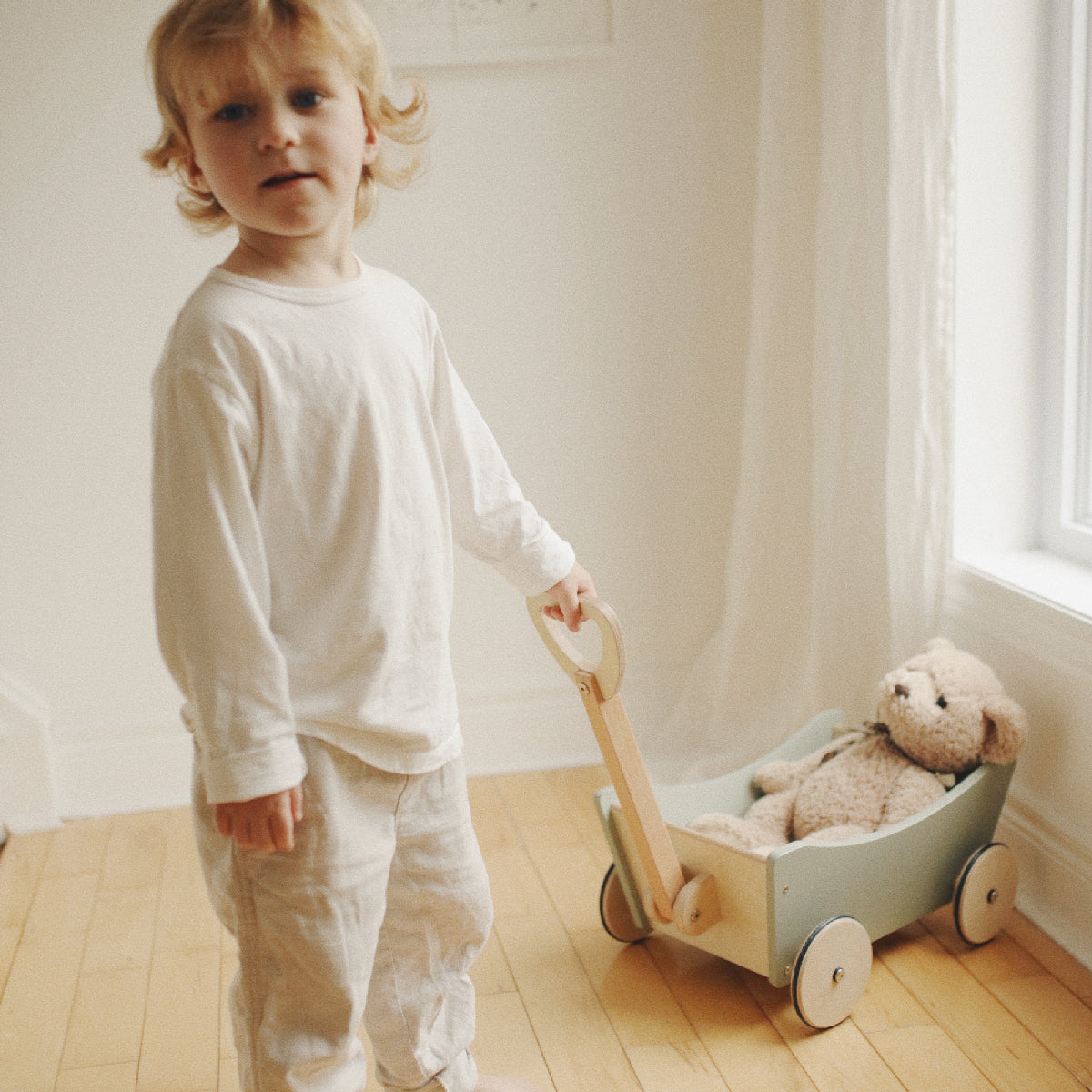Child in white pajamas pushing a wooden toy cart with a teddy bear inside.