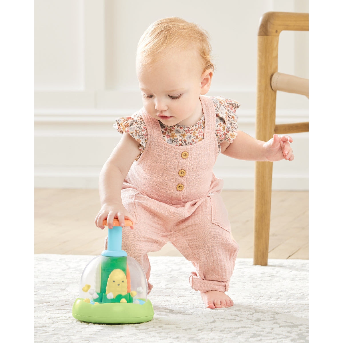 Baby in a pink outfit playing with a green toy on a light-colored floor.